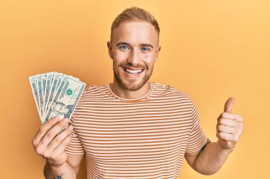 Elinde dolar tutan beyaz bir adam mutlu ve pozitif gülümsüyor, başparmağıyla mükemmel ve onay işareti yapıyor. 