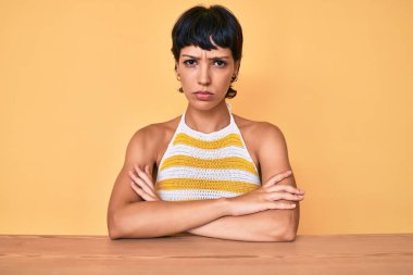 Masada oturan güzel esmer kadın sarı arka plan kuşkucu ve gergin yüzünde çapraz kollarla onaylamayan ifade. negatif kişi. 