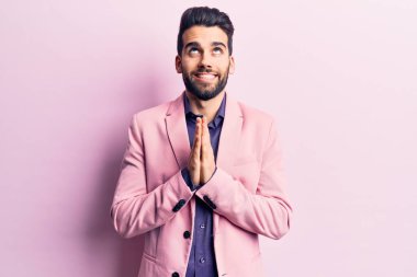 Sakallı, zarif ceketli genç yakışıklı adam elleri bağlı yalvarıyor ve dua ediyor. Yüzünde umut ifadesi var. Çok duygusal ve endişeli. Yalvarıyor. 