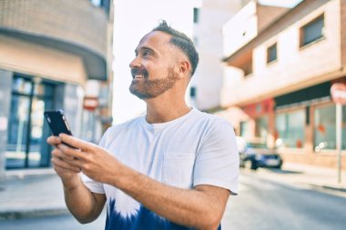 Orta yaşlı yakışıklı adam şehirde akıllı telefon kullanarak mutlu gülümsüyor..