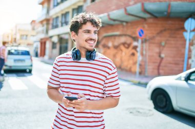 Beyaz adam mutlu bir şekilde gülümsüyor. Kulaklık takıyor, akıllı telefon kullanıyor ve şehirde yürüyor..