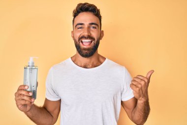 Genç İspanyol adam el dezenfektanı jeli kullanıyor baş parmağını yana uzatıyor ve ağzı açık gülümsüyor. 