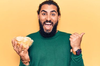Genç Arap adam patates cipsi tutuyor başparmağını yana doğru uzatıyor ve ağzı açık gülümsüyor. 