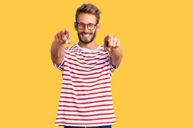 Sakallı, sıradan kıyafetler giyen ve gözlüklü yakışıklı sarışın adam seni ve kamerayı parmaklarıyla gösteriyor, gülümsüyor ve neşeli. 