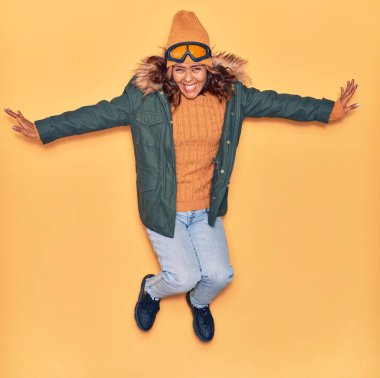 Kışlık giysiler giyen genç, güzel Latin kadın mutlu gülümsüyor. Yüzünde ve kollarında tebessümle, izole edilmiş sarı arka planda atlıyor.