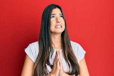 Genç esmer kadın, kırmızı arka plan üzerine günlük beyaz tişört giyip elleri bağlı yalvarıyor ve dua ediyor. Yüzünde umut ifadesi var. Çok duygusal ve endişeli. Yalvarıyor. 