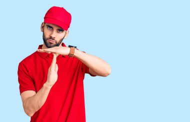 Sakallı, teslimat üniformalı genç yakışıklı adam. Elleriyle jest yapıyor, hayal kırıklığına uğramış ve ciddi bir yüz ifadesi var. 