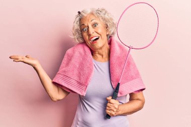 Havlu giymiş, elinde badminton raketi tutan yaşlı, kır saçlı bir kadın başarıyı mutlu bir gülümsemeyle kutluyor ve kazanan el kaldırarak ifade ediyor. 