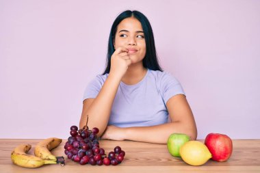 Genç ve güzel Asyalı kız taze ve sağlıklı meyve yiyor. Kuşkuya odaklanmış, çenesinde parmak olan ve kafasını kaldırıp merak eden biri. 