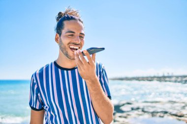Genç İspanyol adam gülümsüyor. Kumsalda sesli mesajla akıllı telefondan konuşuyor..
