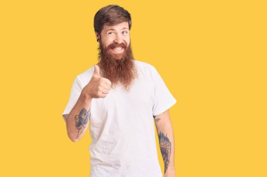 Uzun sakallı, sıradan beyaz tişörtlü, yakışıklı, genç, kızıl saçlı bir adam. Elle mutlu bir jest yapıyor. Kameraya bakıp başarıyı gösteren ifade. 