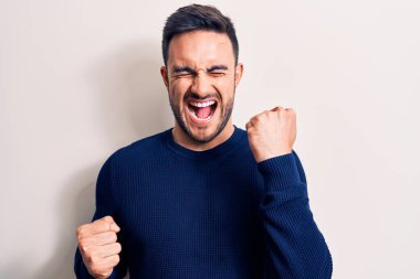 Sakallı, sıradan bir süveter giyen genç, beyaz arka plan üzerinde duran yakışıklı adam, kolları havada ve gözleri kapalı bir şekilde başarıyı kutluyor ve hayrete düşüyor.