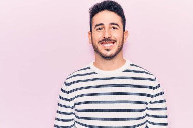 Gündelik kıyafetler giyen genç İspanyol adam pozitif ve mutlu görünüyor. Kendine güvenen bir gülümsemeyle ayakta duruyor ve dişlerini gösteriyor. 