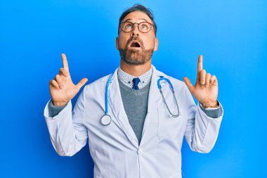 Doktor üniforması ve steteskop giyen yakışıklı orta yaşlı bir adam şaşırmış ve şaşırmış bir şekilde yukarı bakıyor ve parmaklarıyla işaret ediyor ve kollarını kaldırıyor.. 