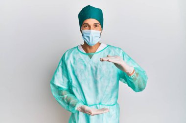 Cerrah üniforması giyen genç yakışıklı bir adam ve elleri büyük ve büyük bir işaret gösteren tıbbi maske, ölçü sembolü. Kameraya bakıp gülümsüyor. ölçüm kavramı. 