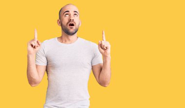 Gündelik tişört giyen genç yakışıklı şaşırmış ve şaşırmış bir şekilde yukarı bakıyor ve parmaklarıyla işaret ediyor ve kollarını kaldırıyor.. 