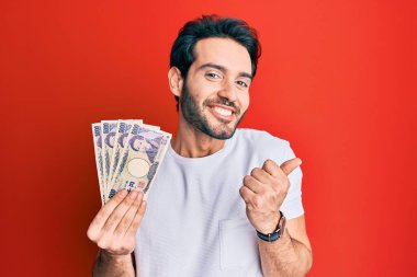 Japon yen banknotları tutan İspanyol genç adam gururla bağırıyor, zaferi kutluyor ve başarıyı kollarını kaldırarak kutluyor. 