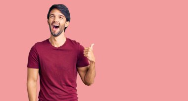 Yakışıklı İspanyol bir adam, günlük kıyafetler giyiyor, gülümsüyor, gülümsüyor ve başparmağıyla yan tarafı işaret ediyor.. 