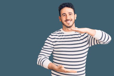 Latin asıllı genç adam, sıradan kıyafetler giyiyor. Elleriyle işaret ediyor. Ölçü sembolü, büyük boy işareti. Kameraya bakıp gülümsüyor. ölçüm kavramı. 