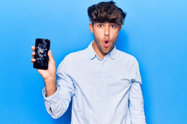 Kırık akıllı telefon tutan genç İspanyol adam kırık ekranı gösteriyor korkmuş ve ağzı açık şaşkınlık içinde, inançsız yüz. 