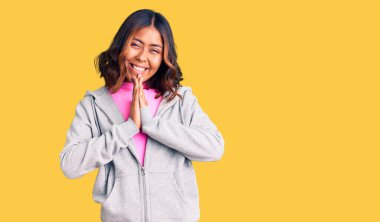 Genç, güzel, melez, sportif kıyafetler giyen, elleriyle dua eden, affedilmek isteyen, kendinden emin, gülümseyen bir kadın.. 