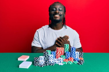 Poker fişleri ve kartlarla masada oturan yakışıklı genç bir adam elleri göğsünde, gözleri kapalı ve yüzünde minnet dolu bir jest ile gülümsüyor. sağlık kavramı. 