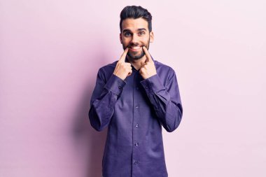 Sakallı, sıradan tişörtlü, ağzı açık gülümseyen, parmakları işaret eden ve neşeli gülümsemeye zorlayan genç yakışıklı bir adam. 