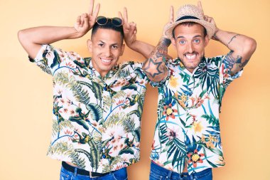 Genç eşcinsel iki adam yaz şapkası takıyor ve Hawaii gömleği giyiyor komik ve çılgın pozlar veriyor parmakları kafasında tavşan kulağı, neşeli bir gülümseme 