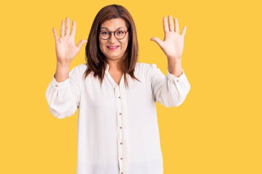 Orta yaşlı Latin kadın gündelik giysiler ve gözlük takıyor ve parmaklarını on numara gösteriyor ve gülümsüyor kendinden emin ve mutlu. 