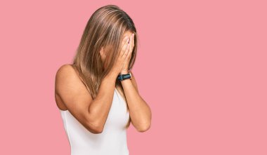 Kolsuz tişörtlü, sıradan giyinen genç, sarışın bir kadın. Ağlarken yüzünü ellerle kapatıyor. depresyon kavramı. 