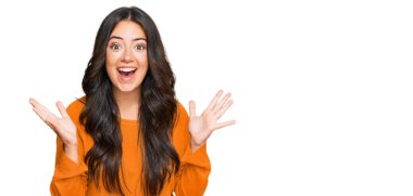 Güzel esmer, turuncu süveter giyen genç bir kadın zaferi mutlu bir gülümsemeyle ve ellerini kaldırarak kazanan bir ifadeyle kutluyor. 