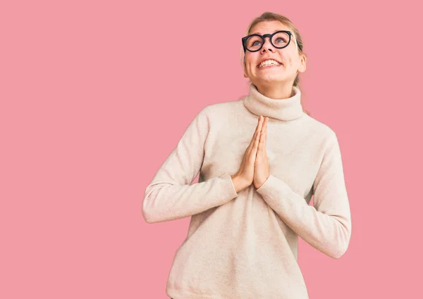 Young beautiful blonde woman wearing turtleneck sweater and glasses begging and praying with hands together with hope expression on face very emotional and worried. begging.