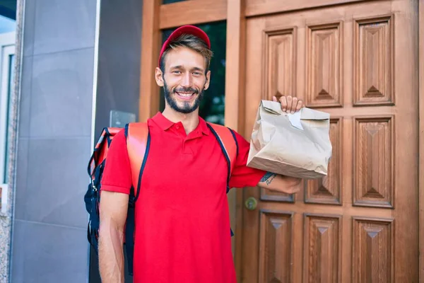 赤い制服と配送バックパックを身に着けている白人の配達人笑顔幸せな屋外保持紙バッグ — ストック写真