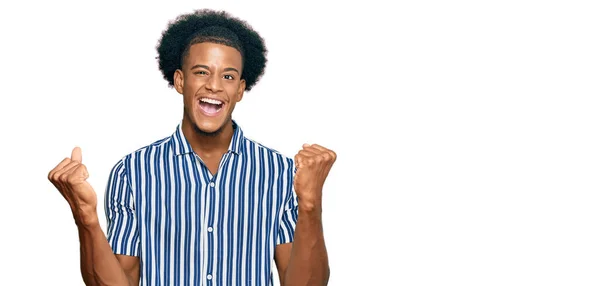 アフロの髪をしたアフリカ系アメリカ人の男性は カジュアルな服を着て非常に満足し 腕を上げ 笑顔と成功のために叫んで勝者のジェスチャーを行うことに興奮しています お祝いのコンセプト — ストック写真