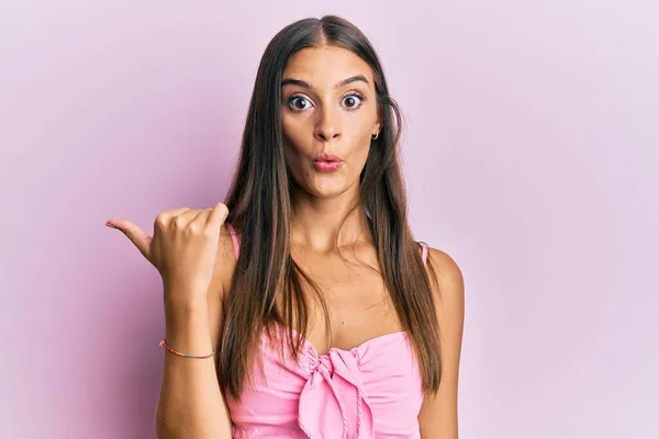 Young hispanic woman wearing casual style with sleeveless shirt surprised pointing with hand finger to the side, open mouth amazed expression.