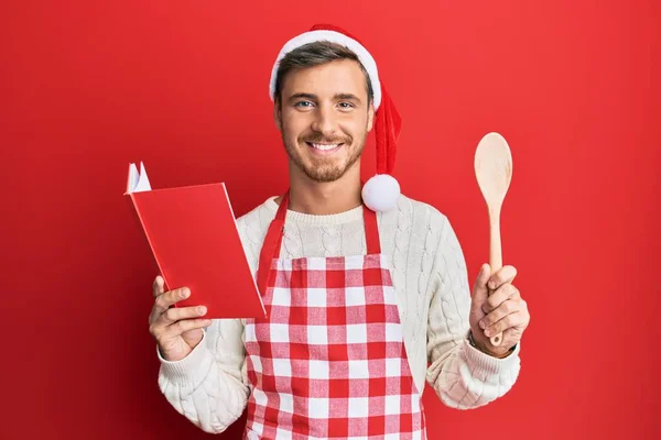 英俊的高加索人穿着烤肉围裙和圣诞帽做菜 脸上挂着快乐而冷静的笑容 显示牙齿 — 图库照片