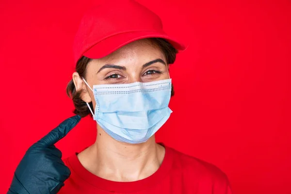 Young hispanic girl wearing delivery uniform and covid-19 safety mask smiling happy pointing with hand and finger