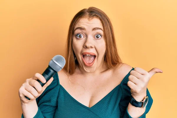 Young Beautiful Redhead Woman Singing Song Using Microphone Pointing Thumb — Stock Photo, Image