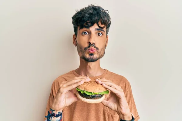 Jovem Hispânico Comendo Saboroso Hambúrguer Clássico Olhando Para Câmera Soprando — Fotografia de Stock