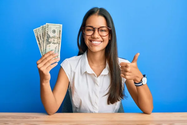 Beautiful hispanic woman holding 20 dollars banknotes smiling happy and positive, thumb up doing excellent and approval sign