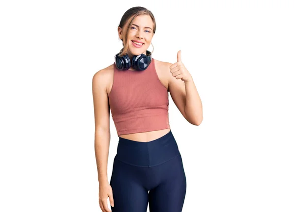 漂亮的高加索年轻女人穿着运动服 用耳机做着快乐的大拇指手势 赞成的表情看着相机显示的成功 — 图库照片