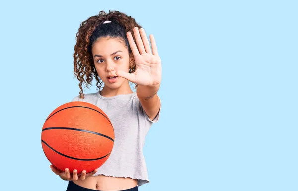 美しいです子供の女の子とともに巻き毛保持バスケットボールボールでオープン手を行います停止標識で真剣で自信を持って式 防衛ジェスチャー — ストック写真