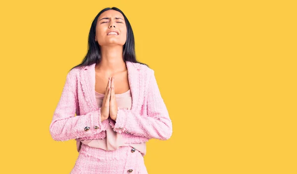 Young beautiful latin girl wearing business clothes begging and praying with hands together with hope expression on face very emotional and worried. begging.