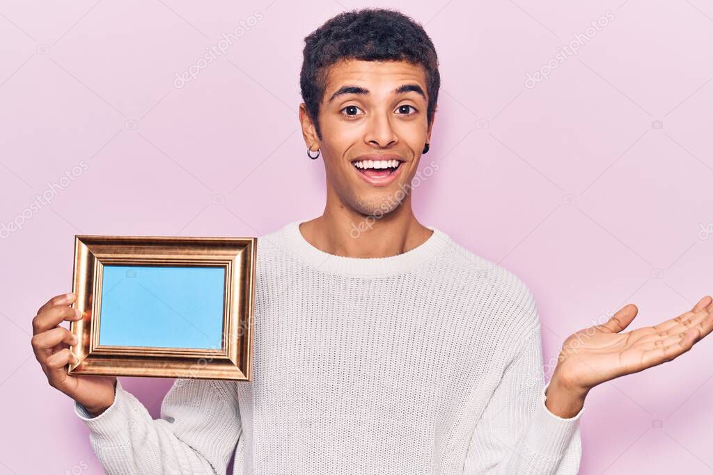 Young african amercian man holding empty frame celebrating achievement with happy smile and winner expression with raised hand 