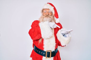 Gri saçlı ve uzun sakallı yaşlı bir adam Noel Baba kostümü giyiyor kameraya hayret ediyor ve gülümsüyor.. 