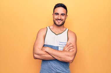 Sarı arka plan üzerinde kolsuz tişört giyen sakallı yakışıklı genç adam kameraya bakarken kollarını kavuşturup gülümsüyor. Pozitif kişi.