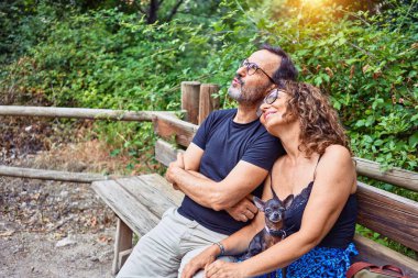 Orta yaşlı çift gökyüzüne bakarken mutlu bir şekilde gülümsüyor. Parkta chihuahua köpeğiyle bankta oturmak.