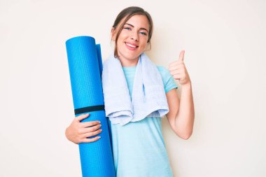 Güzel, beyaz bir kadın yoga minderi tutuyor mutlu ve pozitif gülümsüyor, başparmağıyla mükemmel ve onay işareti yapıyor. 