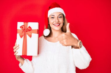 Genç esmer kadın noel şapkası takıyor ve elinde bir hediye tutuyor mutlu ve pozitif gülümsüyor, başparmağıyla mükemmel ve onay işareti yapıyor. 