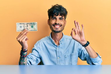 20 dolarlık banknot tutan genç İspanyol adam parmaklarıyla imza atıyor, gülümseyerek mükemmel bir sembol oluşturuyor. 